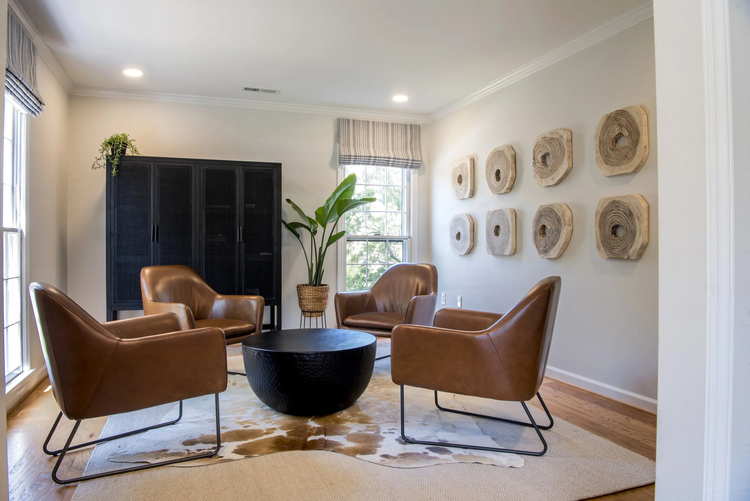 Living room with four brown leather chairs arranged around a black coffee table on a light patterned rug, a black cabinet, a plant in a wicker basket, a window with striped curtains, and wall art made of wooden circles.