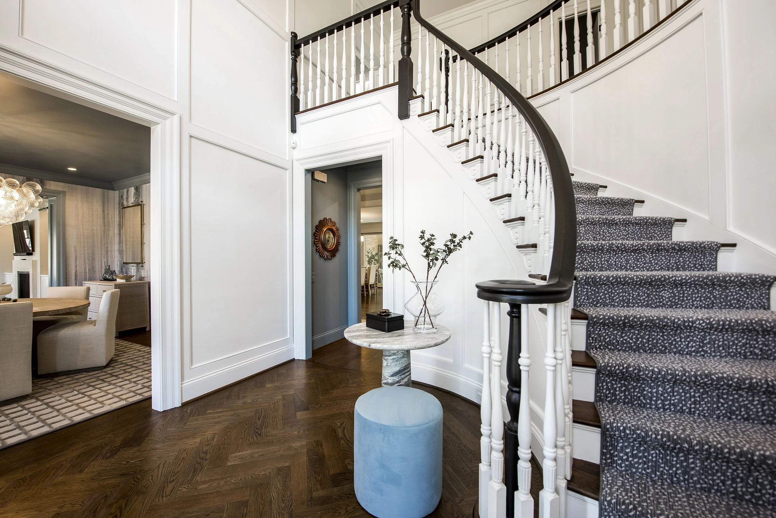 Interior view of a home with a curved staircase carpeted in dark with white speckles, white paneled walls, a small round marble-topped table with a glass vase and decorative branches, a blue ottoman, and a hallway leading to a dining area visible in the background.