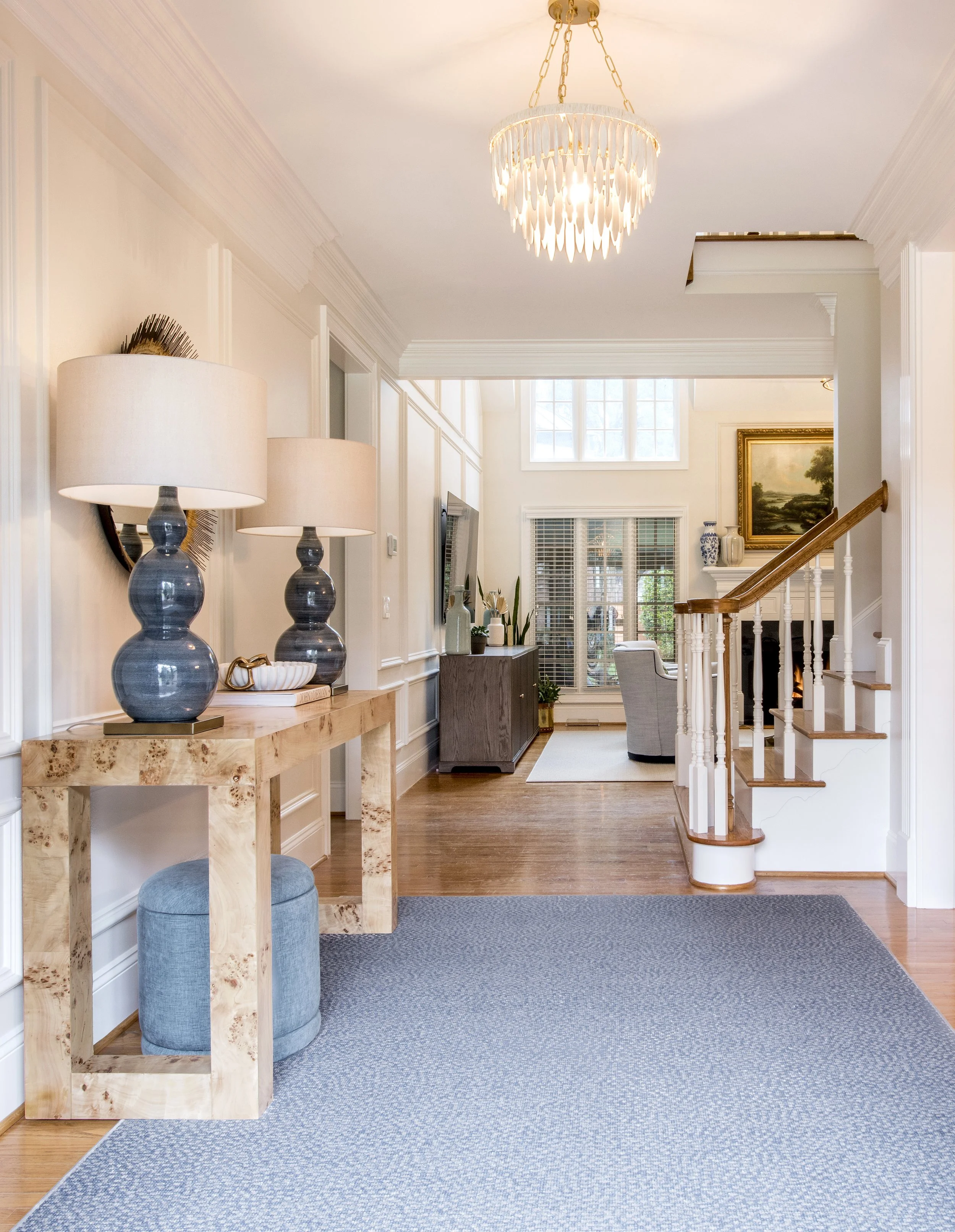 Living room and foyer of a house with a chandelier, lamps, a console table, a rug, a staircase, and furniture including chairs and a cabinet.