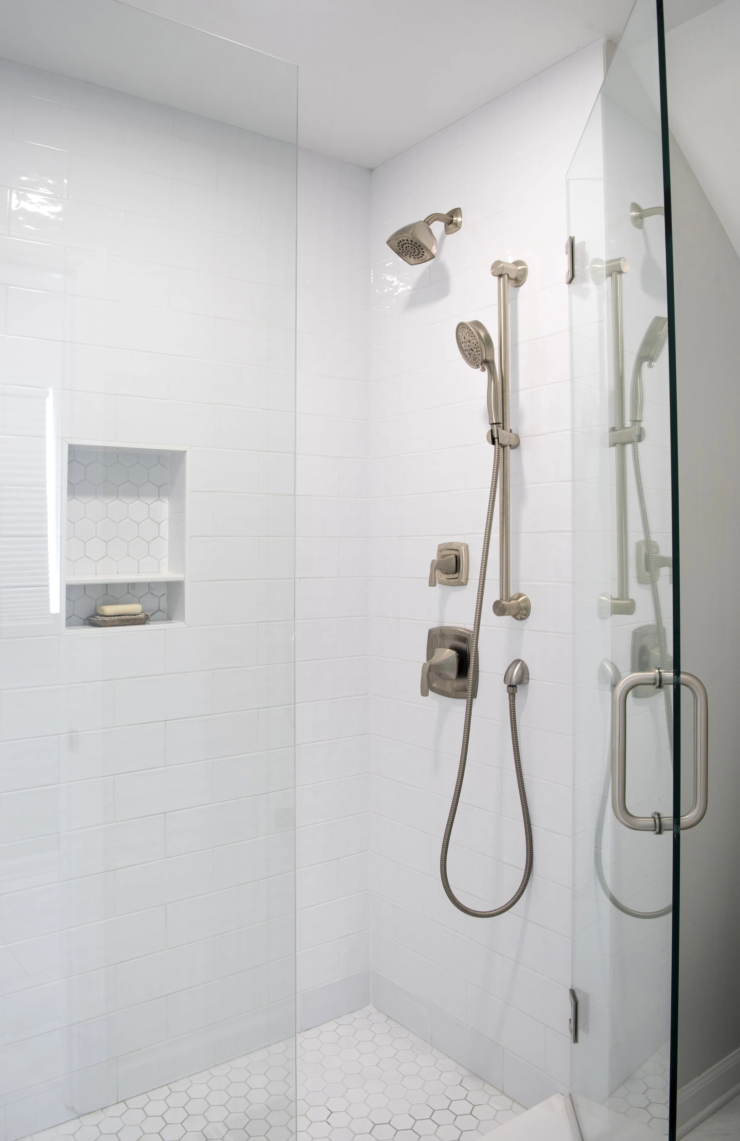 A modern walk-in shower with white tiled walls, a built-in shelf with soap, and adjustable shower heads with stainless steel fixtures.