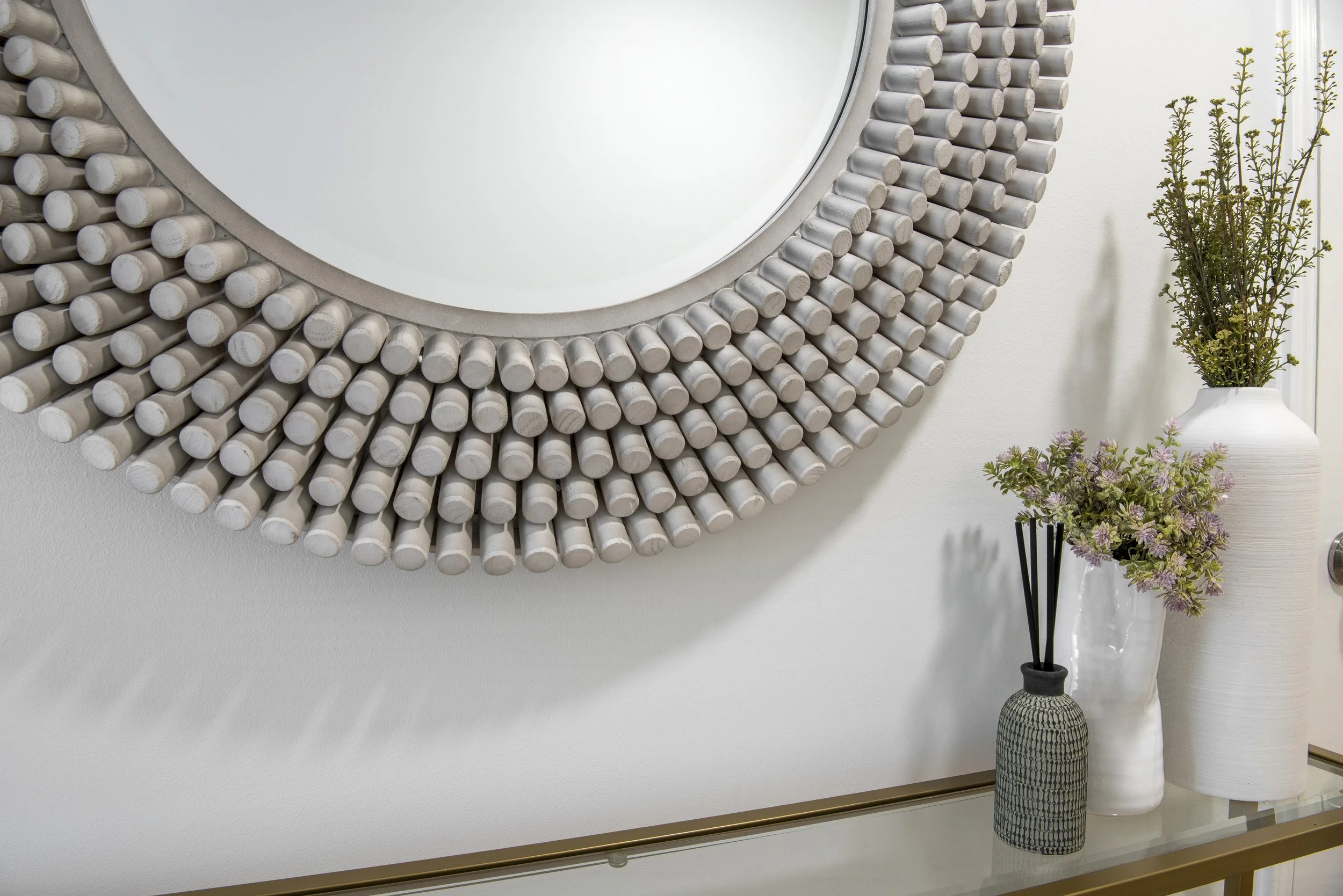 Decorative wall mirror with concentric circular frame made of small, rounded, beige wooden rods, beside a console table with white and gray vases containing tall green plants and purple flowers, and a reed diffuser.