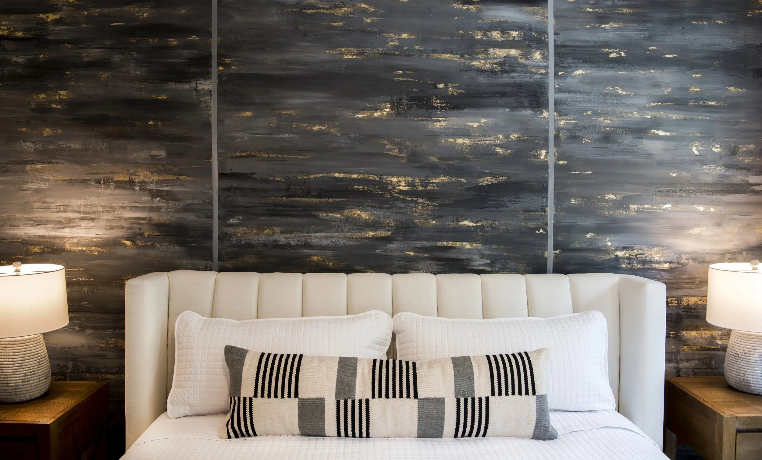 A bedroom scene showing a white upholstered bed with two large white pillows and a decorative rectangular pillow with a black, gray, and white geometric pattern. There are two wooden nightstands, each with a white table lamp with a textured base. The background features a dark, abstract textured wall with streaks of gold and silver accents.