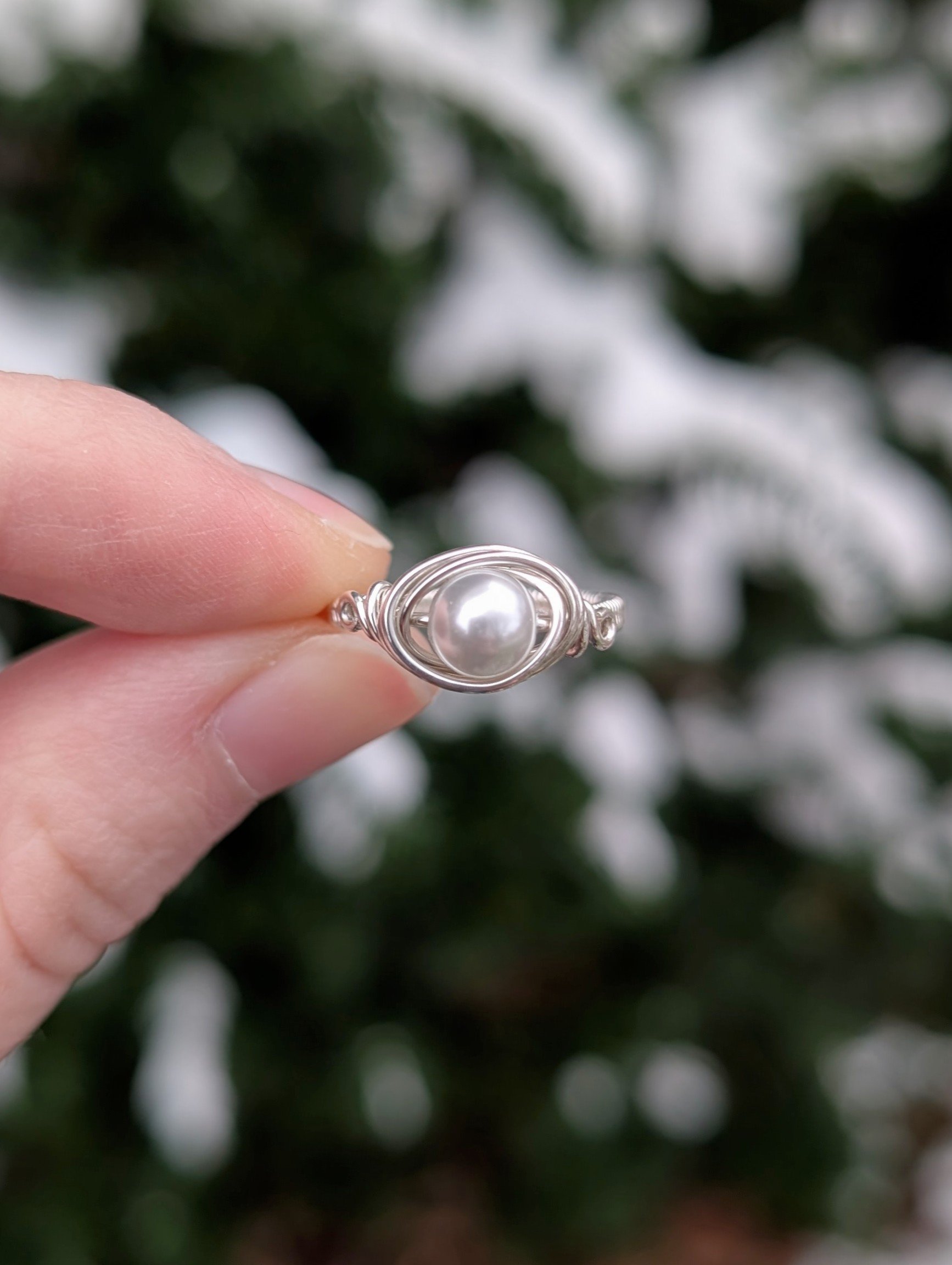 a wire wrapped ring made from a white glass pearl and sterling silver wire held up between two fingers to show the front.