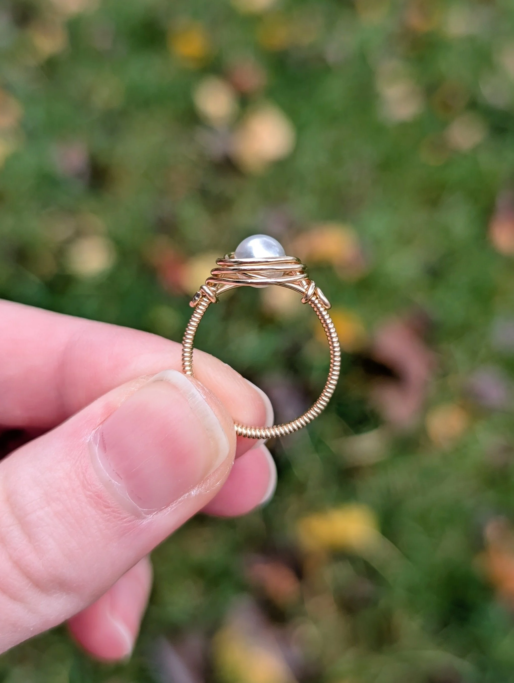 a wire wrapped ring made from a white glass pearl and gold filled wire held up between two fingers to show the side.
