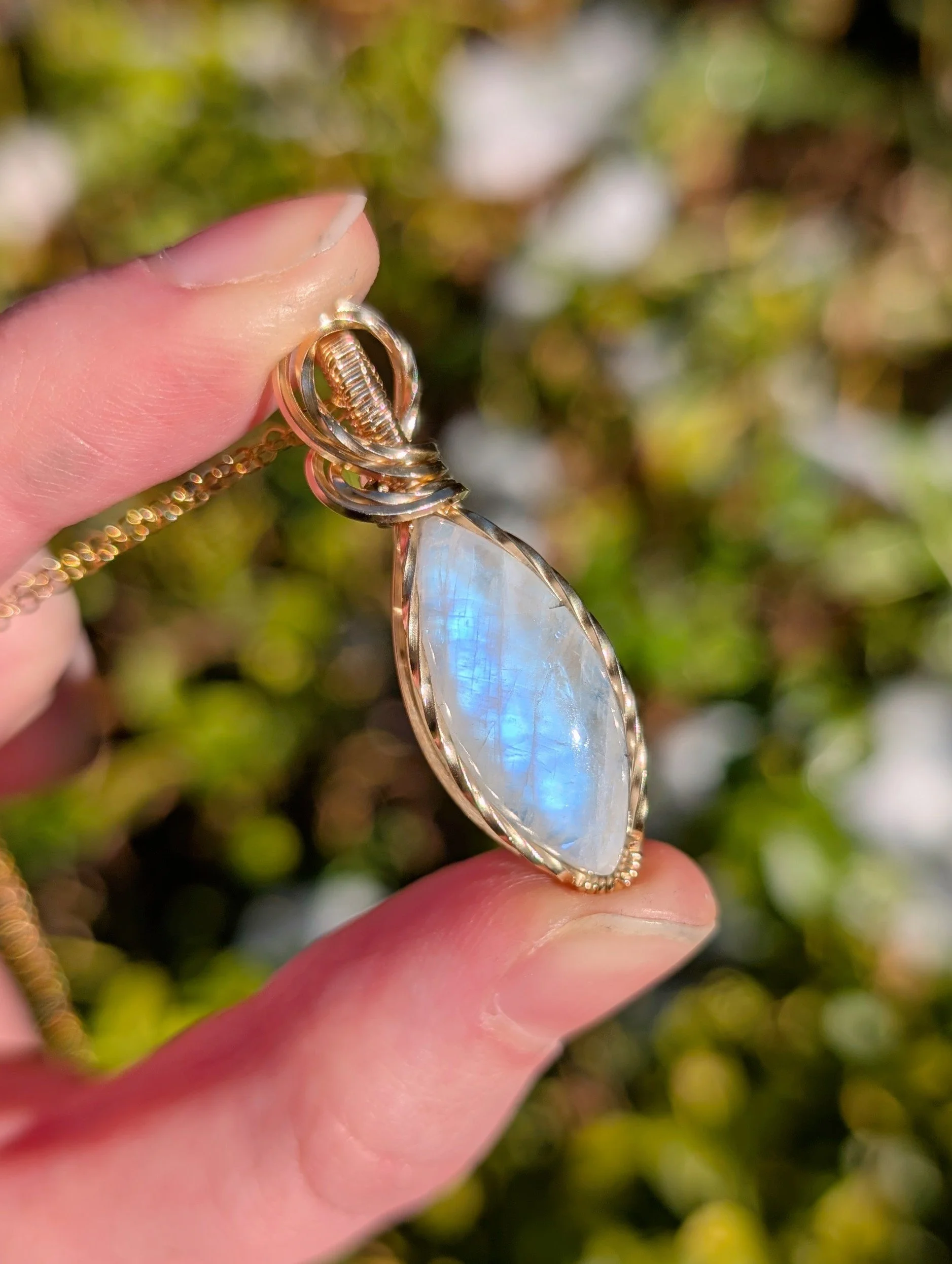 A pendant made with a marqui-shaped moonstone and 14k gold filled wire in direct sunlight. The stone is giving off a soft blue flash.