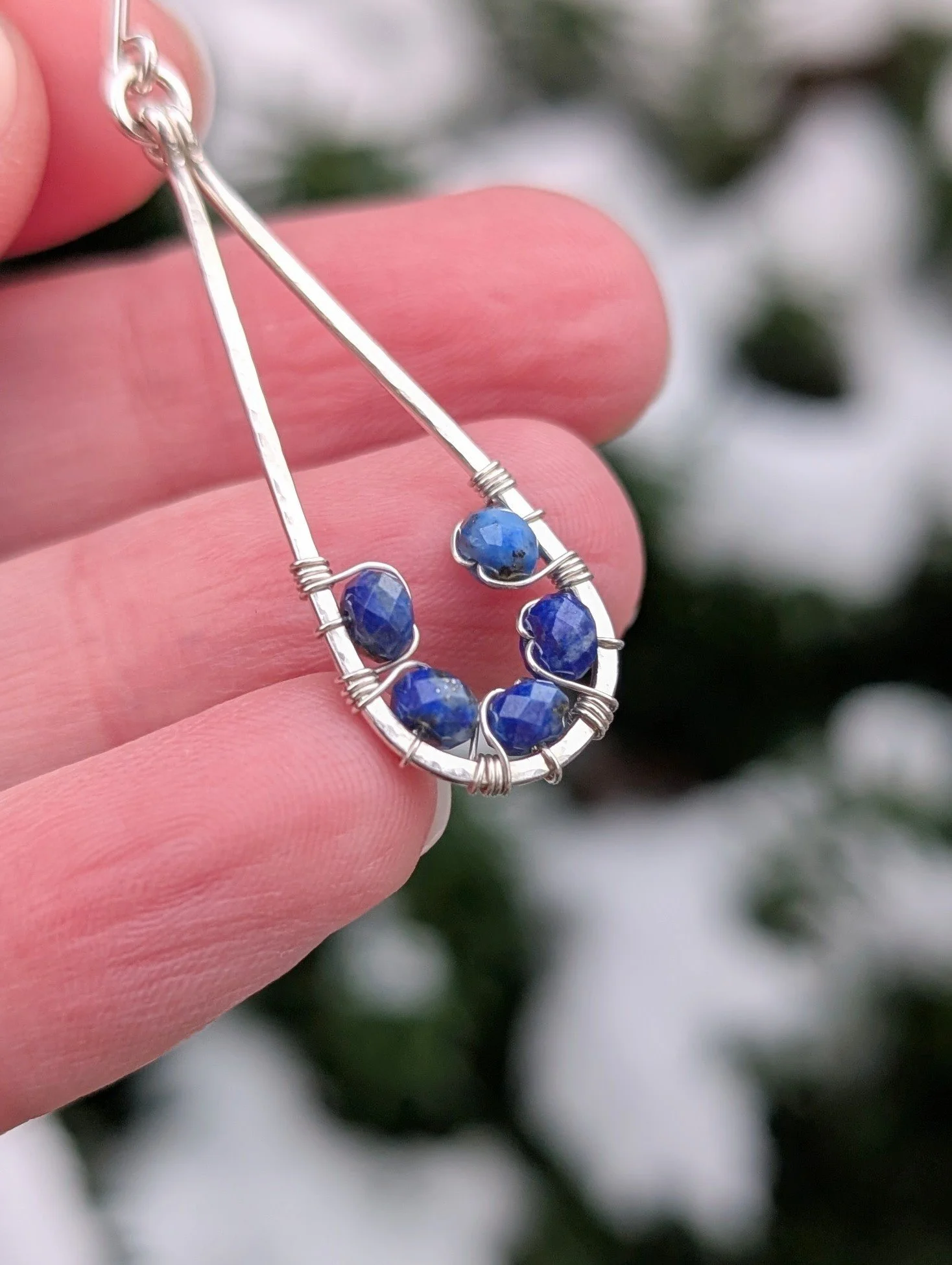 A single Cobalt Sky Earring is being held up close to show the details of the wire wrapping. The earring is made with lapis lazuli beads and sterling silver wire.