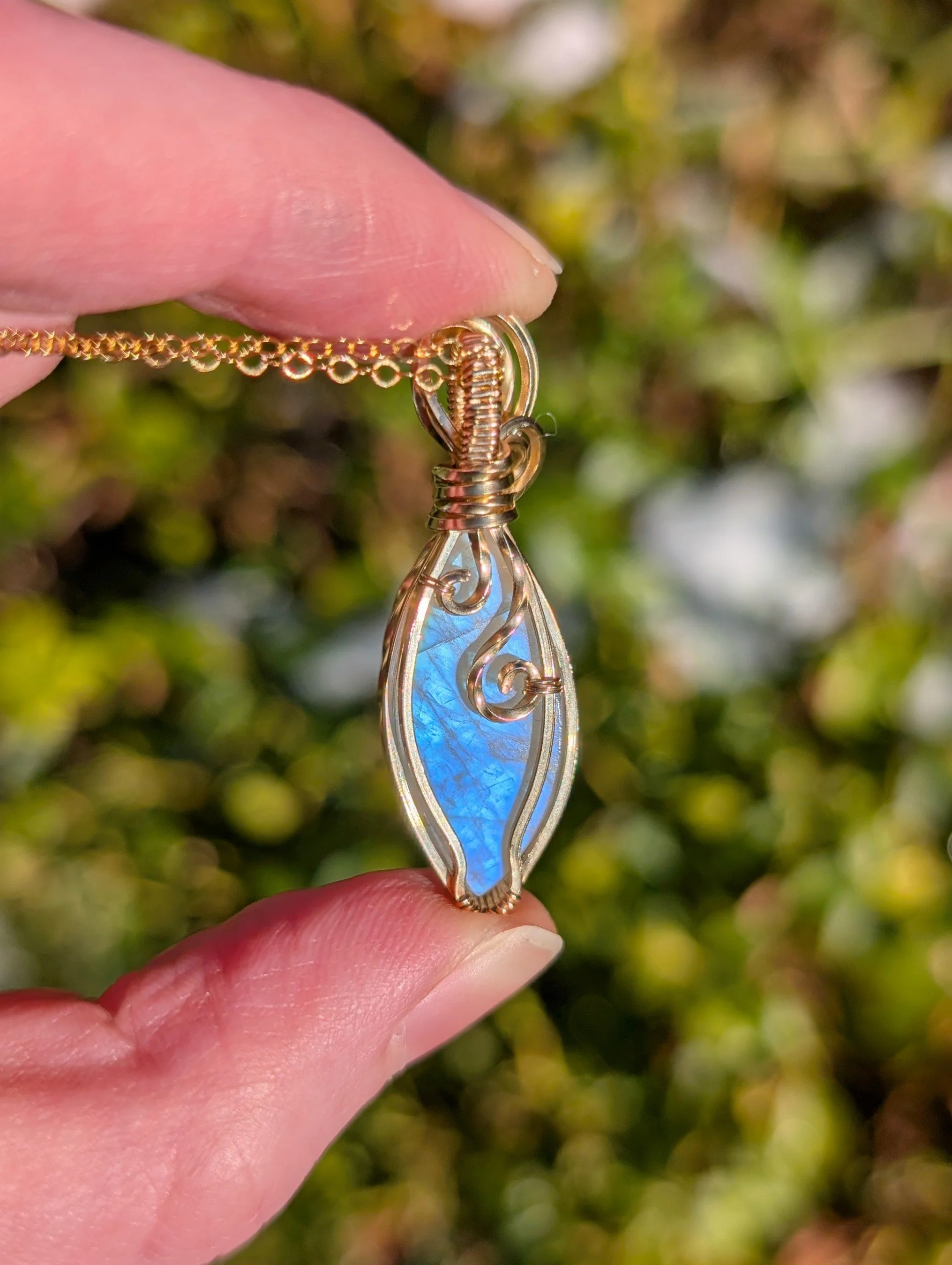 the back of a pendant made with a marqui-shaped moonstone and 14k gold filled wire in direct sunlight. The stone is giving off a soft blue flash.