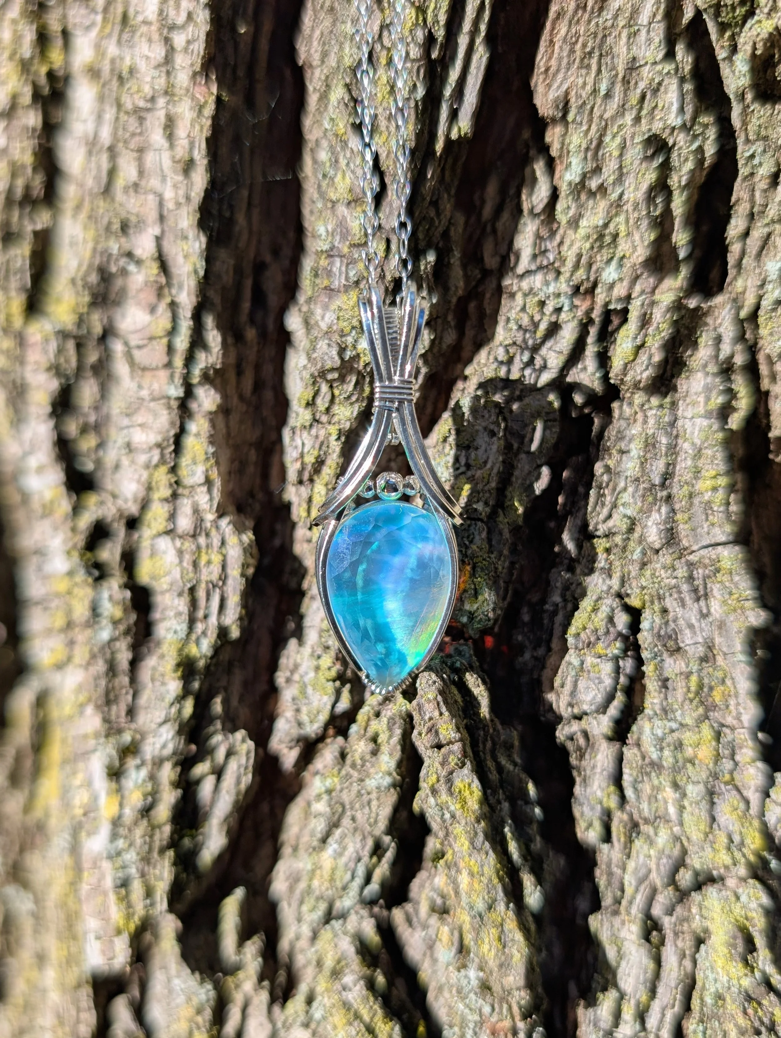 The Vaeloria necklace, featuring a teardrop aurora opal stone wrapped in sterling silver wire. The pendant is propped up against a tree trunk.