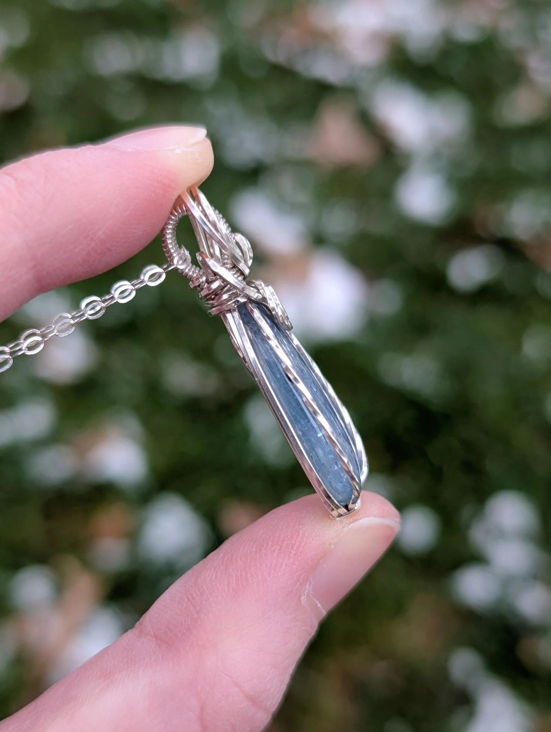 The side of a pendant necklace made from a teardrop shaped icy kyanite stone set in sterling silver wire. It has a sterling silver chain.