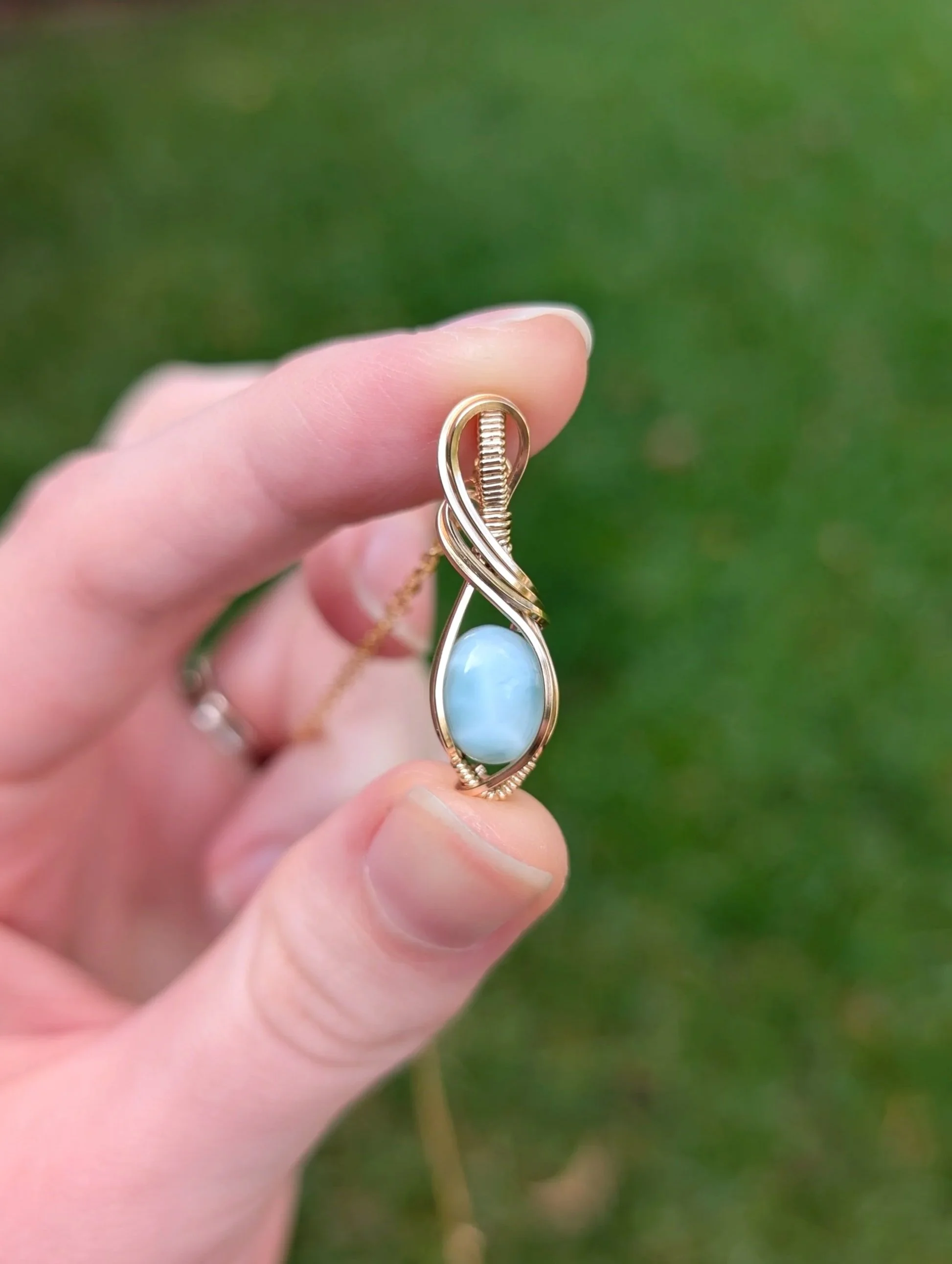 A necklace pendant made from an oval larimar stone and gold filled wire held in a model's hand.