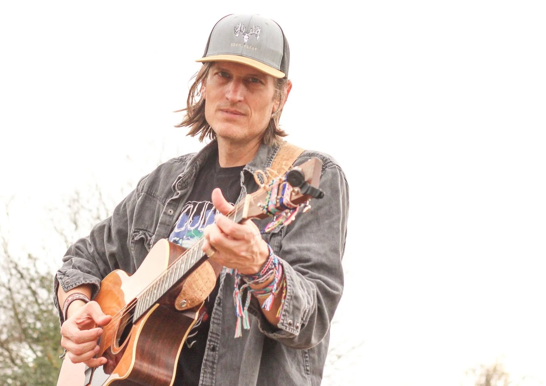 A man with long hair wearing a baseball cap, a black T-shirt with a graphic, and a dark jacket, playing an acoustic guitar outdoors on a bright day.