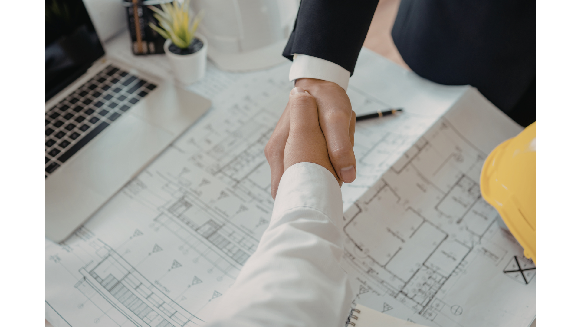 Two people shaking hands over architectural plans on a table, with a laptop, a yellow safety helmet, and a small potted plant nearby.