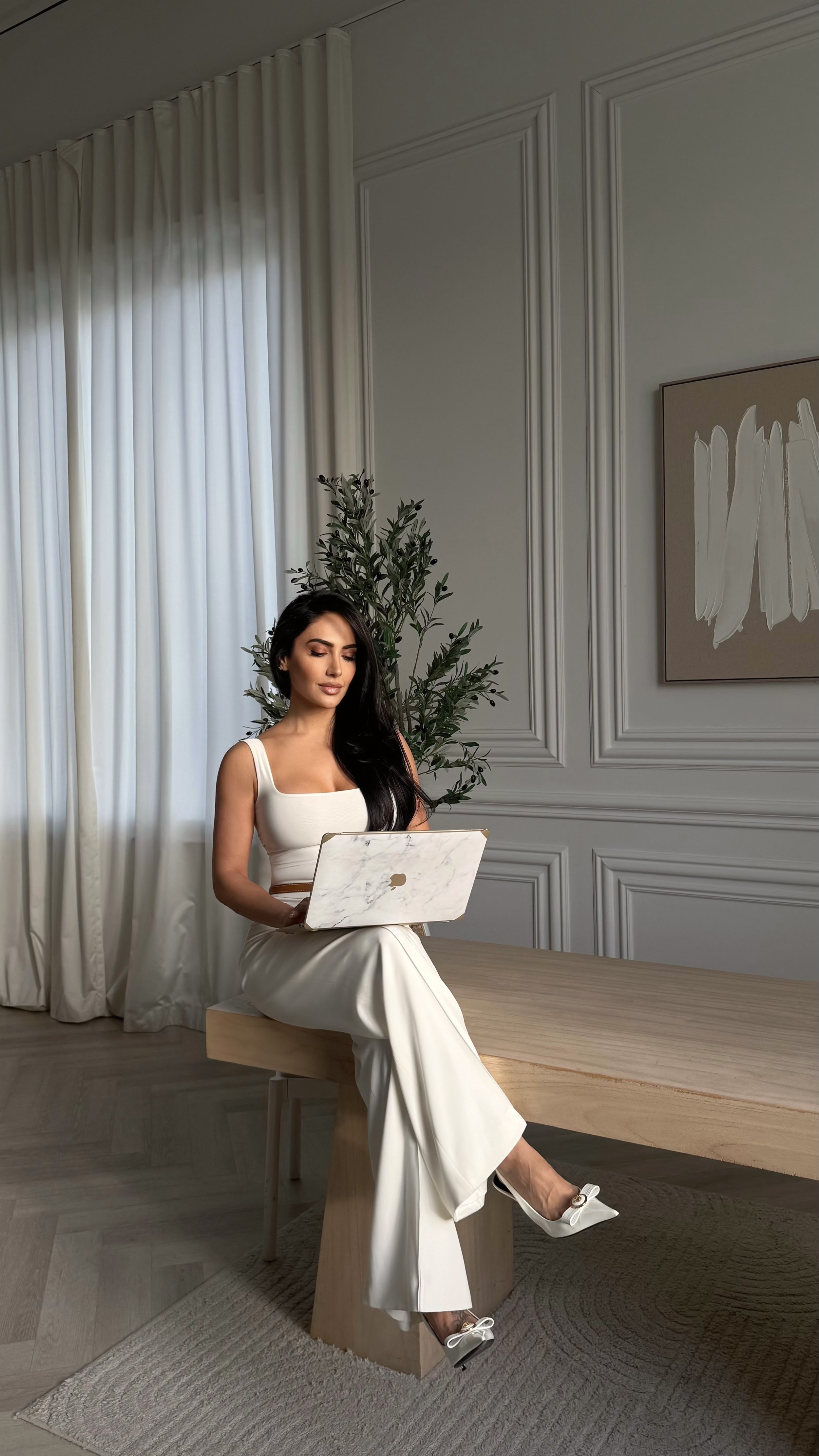 Sydney Sajadi with long dark hair wearing a white outfit sitting on a wooden desk, holding a marble-patterned laptop, in a room with white curtains, white paneled walls, a large painting, and a potted plant.