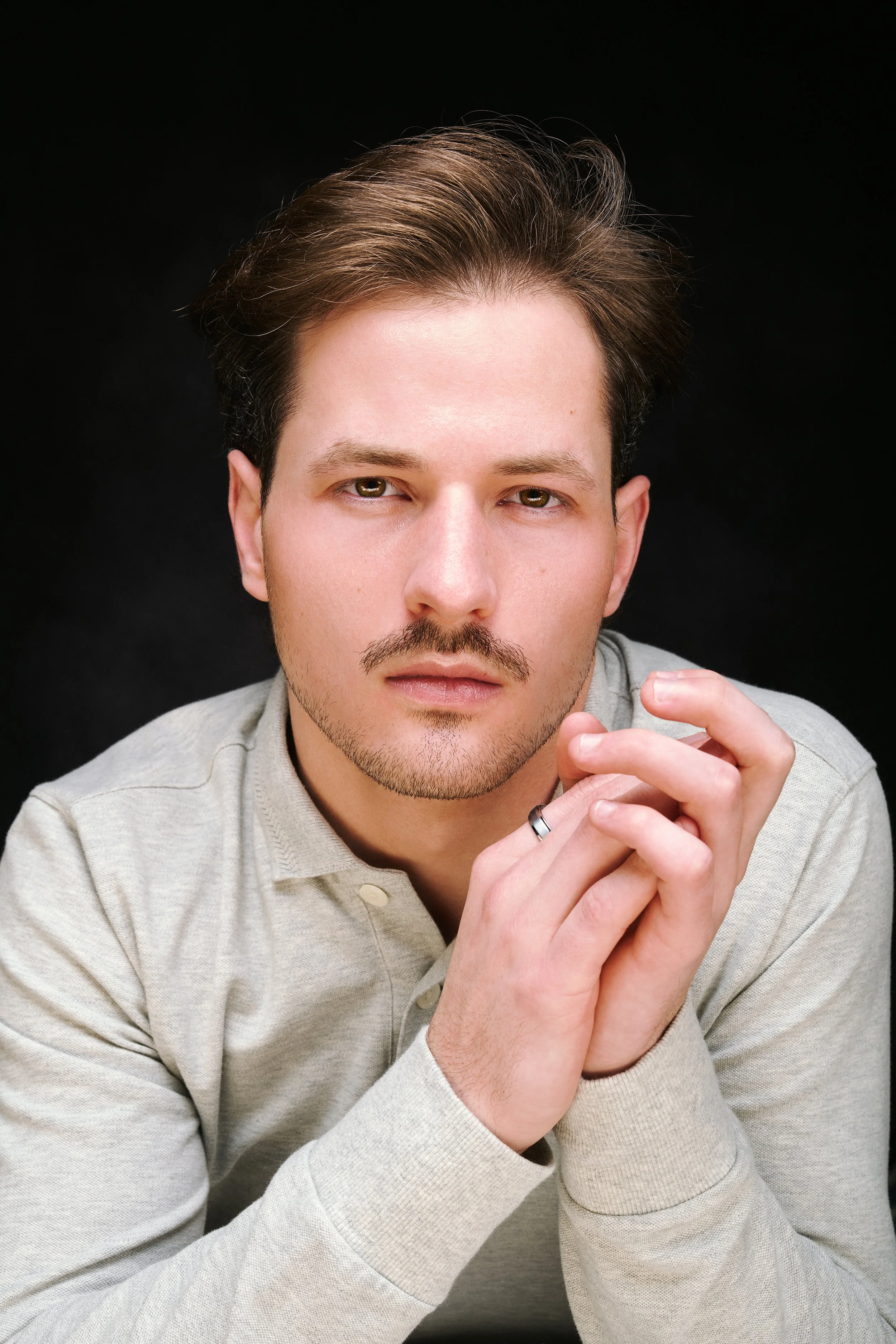 A young man with brown hair and a mustache, wearing a light-colored shirt, looking directly at the camera with his hands clasped together.