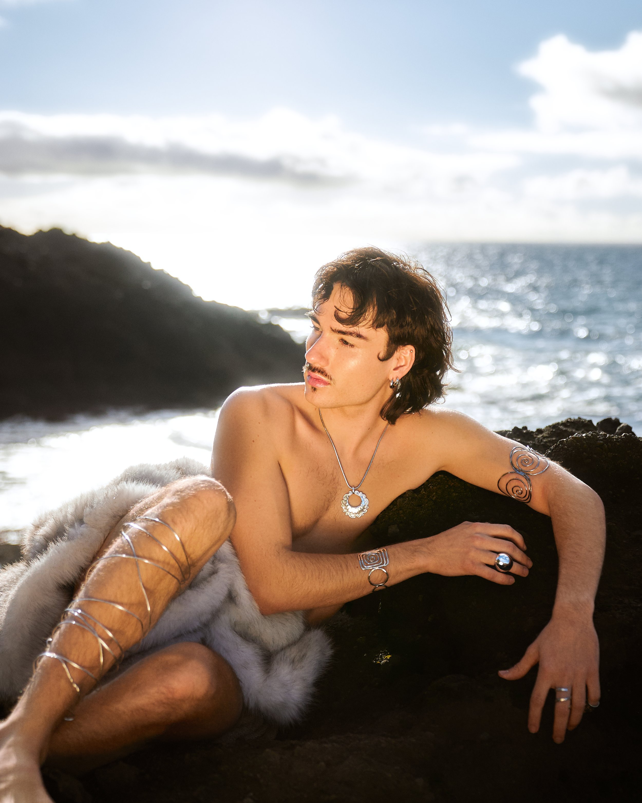 Person sitting on rocks by the ocean, wearing jewelry and a fur-like wrap, with tattoos visible on arm.