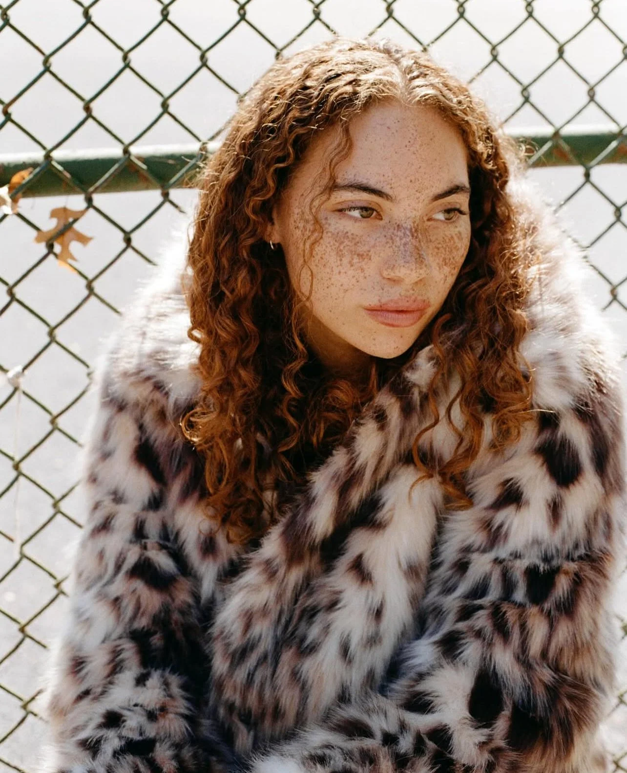 Person with curly hair and freckles wearing a patterned faux fur coat in front of a chain-link fence.