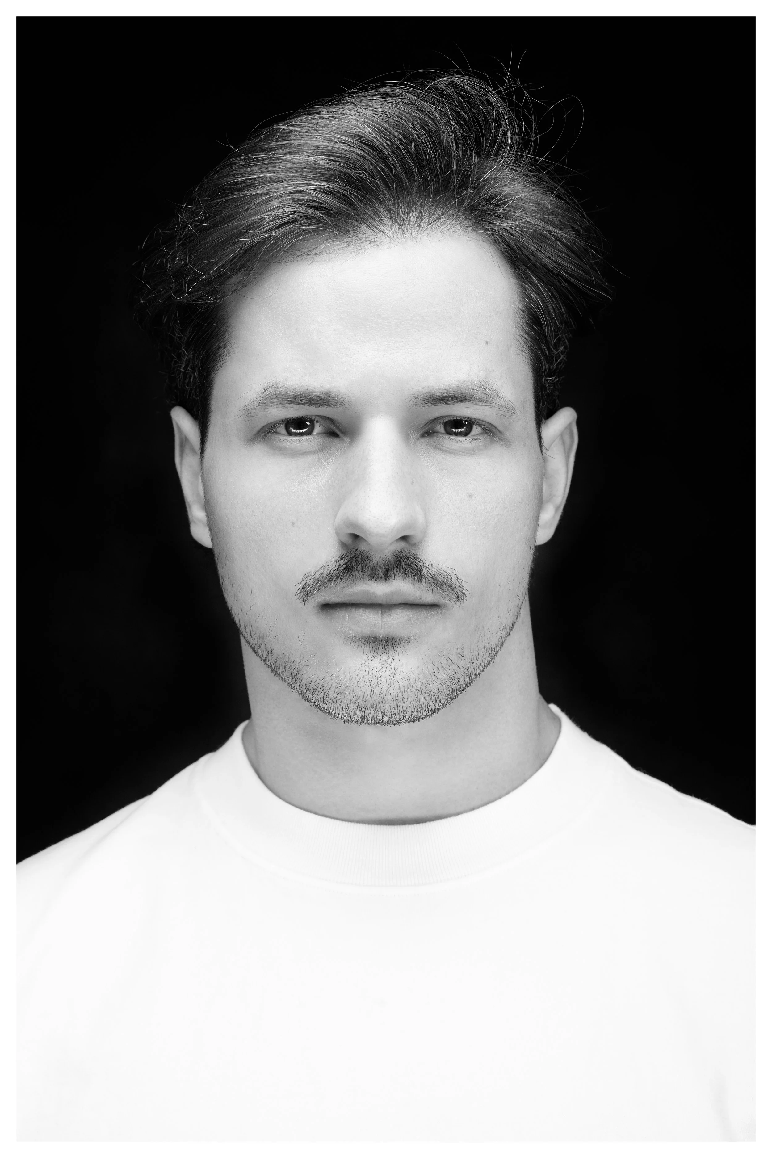 Black and white portrait of a young man with short hair, slight mustache and beard, wearing a white t-shirt, against a dark background.