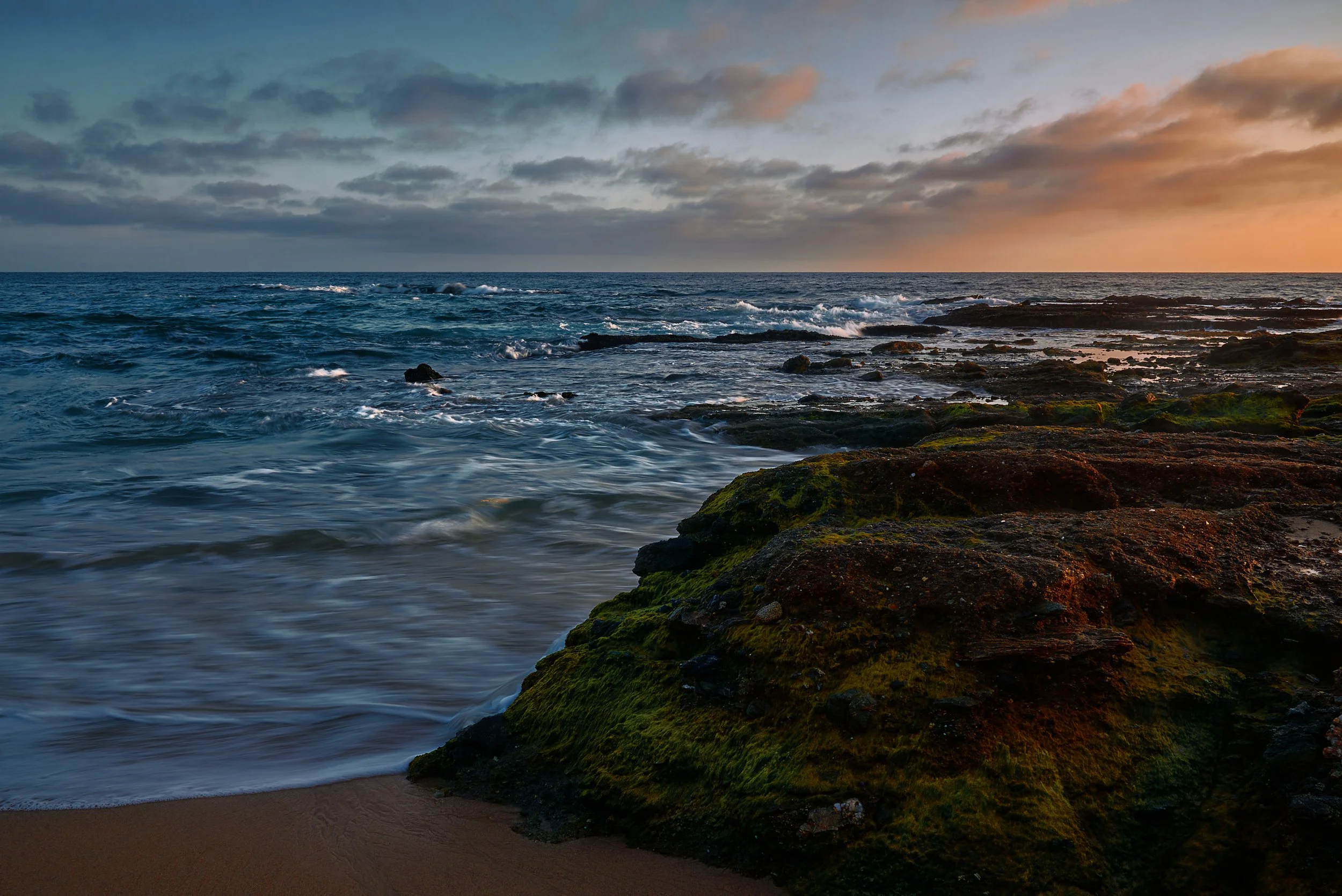 A beach scene at sunset with waves crashing onto the rocky shore covered with green moss and a sandy beach foreground.