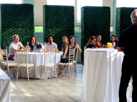 Group of people sitting at round tables with a speaker in a well-lit venue, featuring tall green partitions and drinks on tables.
