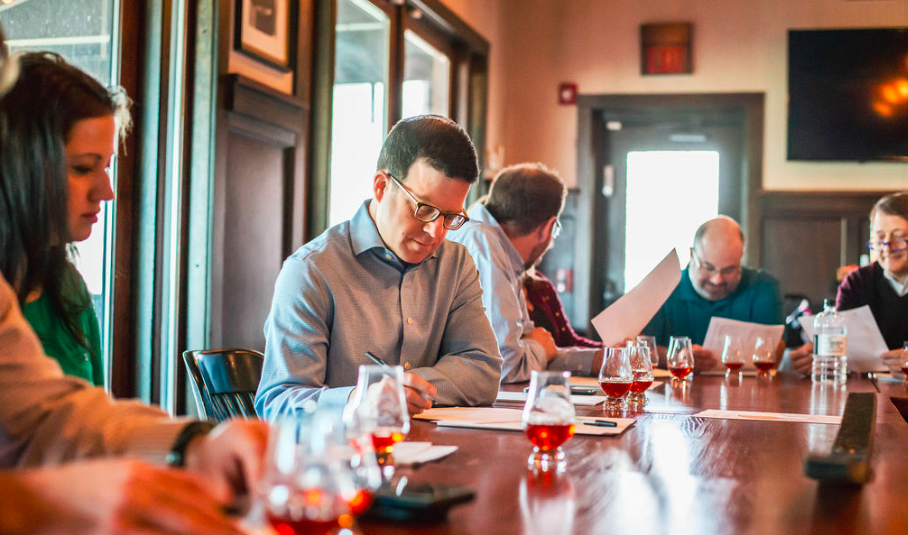 Group of people at a table engaged in a tasting session, with glasses of amber liquid and papers in front of them.