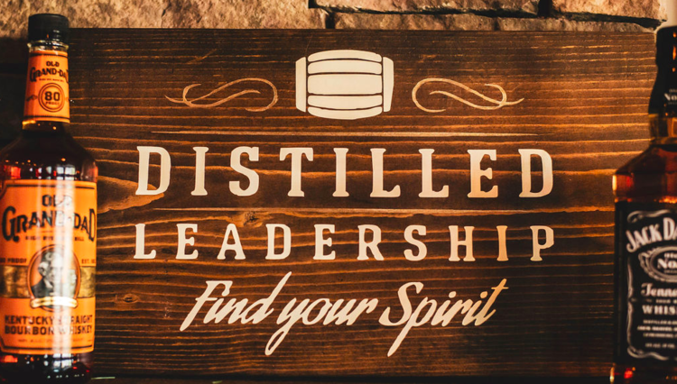 Wooden sign with barrel illustration and text 'Distilled Leadership, Find your Spirit,' flanked by whiskey bottles.