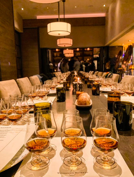 Long table set with multiple glasses of whiskey, candles, menu cards, and a bread basket in an elegantly lit dining room.