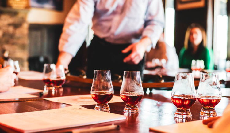 Glasses of whiskey on a wooden table in a bar with people in the background.