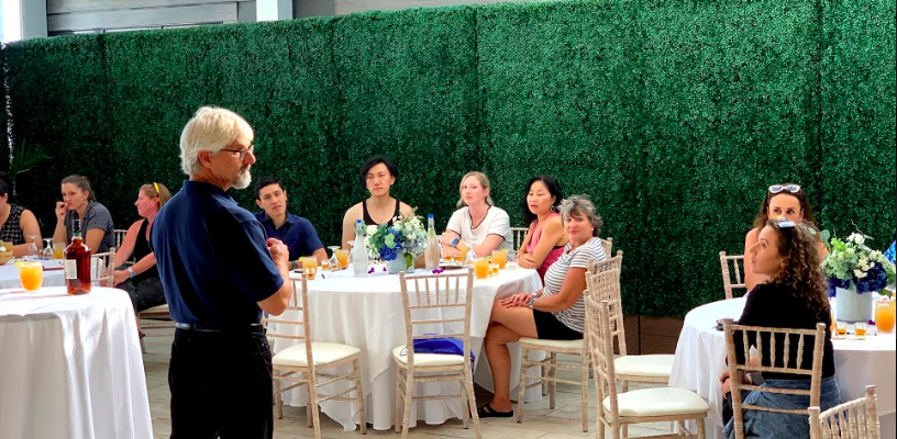 A man speaking to a group of seated people at a gathering with round tables and decorative greenery walls.
