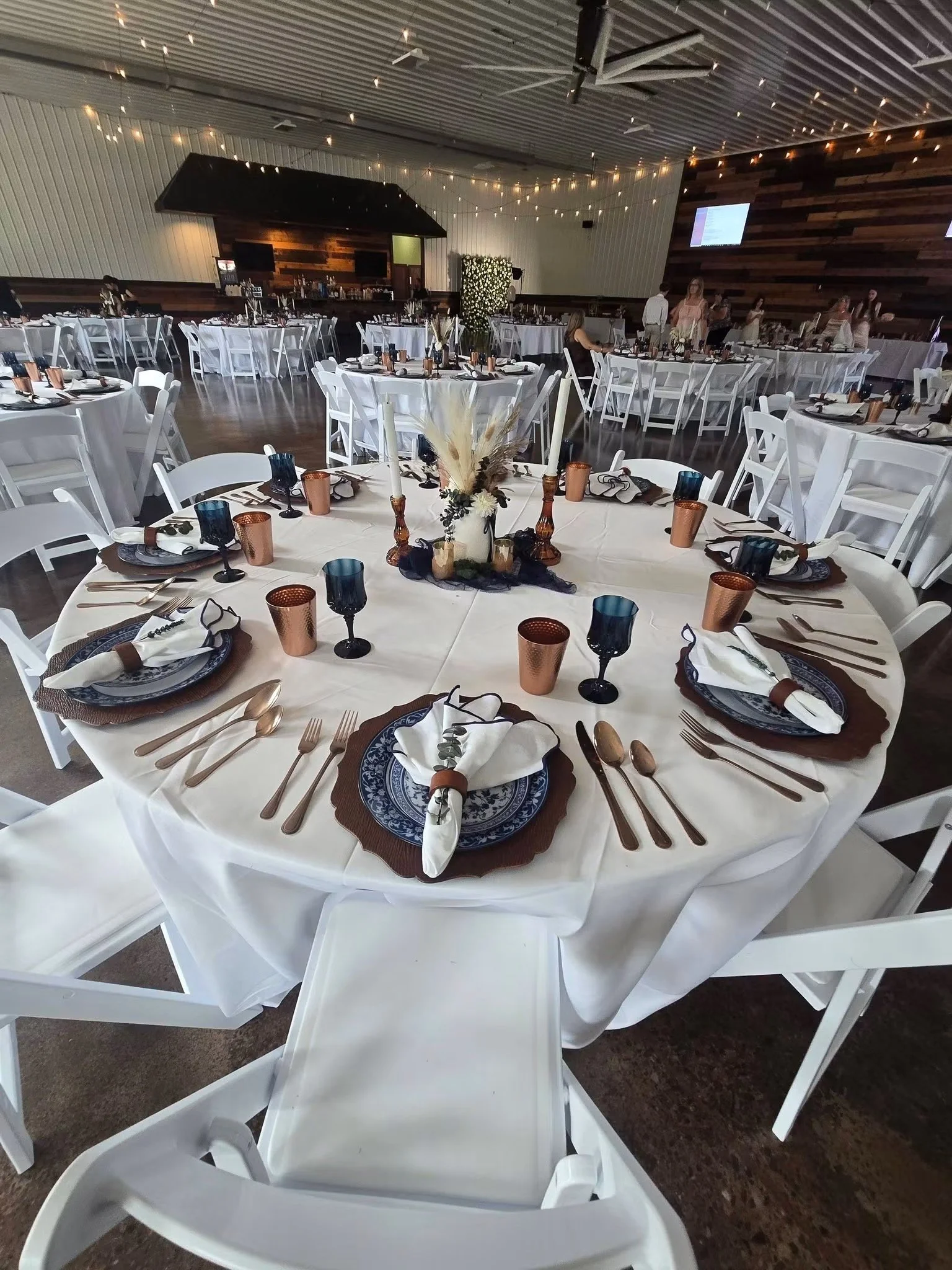 A large banquet hall decorated for an event, with round tables covered in white tablecloths, set with plates, silverware, and glasses. A floral centerpiece with pampas grass and flowers is on the foreground table. The hall has string lights hanging from the ceiling and a wooden accent wall with mounted TV screens.