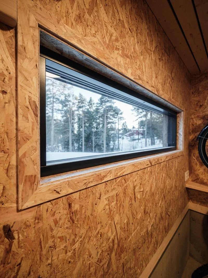 A rectangular window in an unfinished wall with exposed OSB plywood, showing a snowy landscape with trees and a house outside.