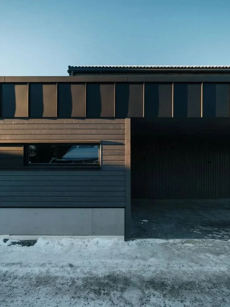 Modern house with dark exterior panels, a window, and a garage or covered parking area, with snow on the ground under a clear blue sky.