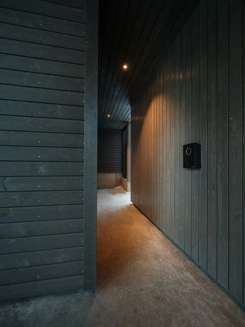 A narrow hallway with dark wood paneled walls and ceiling, illuminated by small ceiling lights, and a wall-mounted speaker on the right.