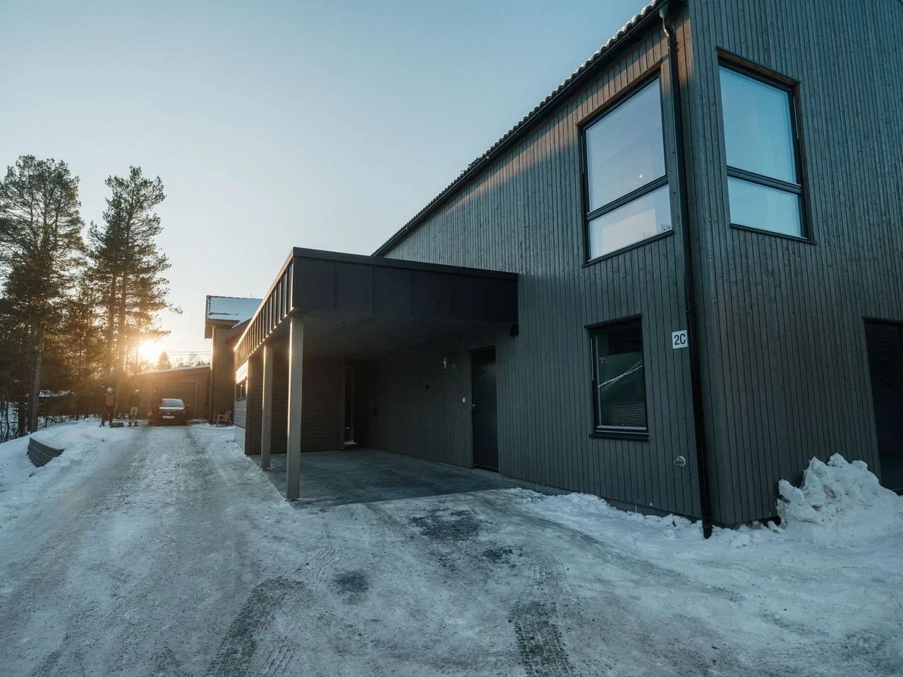 A modern two-story house with black wooden siding and large windows, situated in a snowy environment during sunset.
