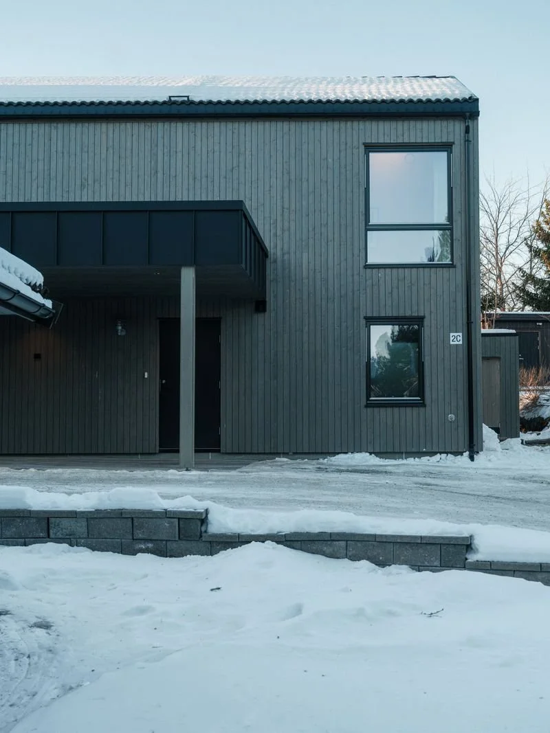 Modern two-story house with dark gray vertical wooden siding, large windows, located in a snowy landscape, with stairs leading up to the entrance.