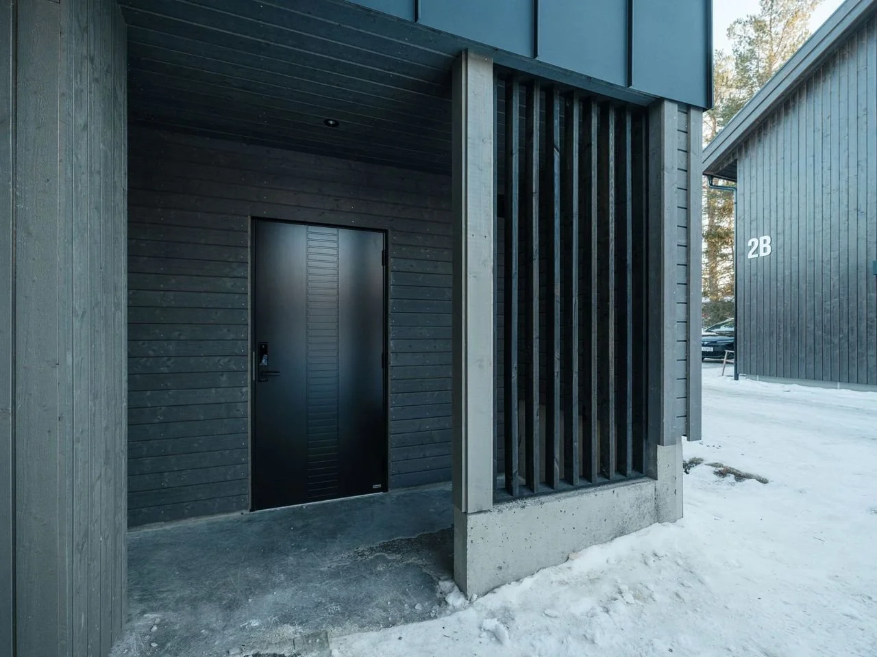 Black front door of a modern building with black wood siding and vertical wooden slats on concrete base. Snow on ground, with neighboring building marked '2B' and parking lot visible in the background.