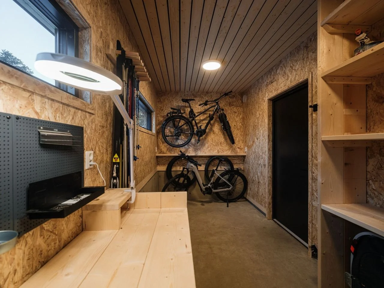 A wooden workshop or storage room with two bikes mounted on the wall, shelves, a pegboard with tools, and a workbench. There are windows and a ceiling light.
