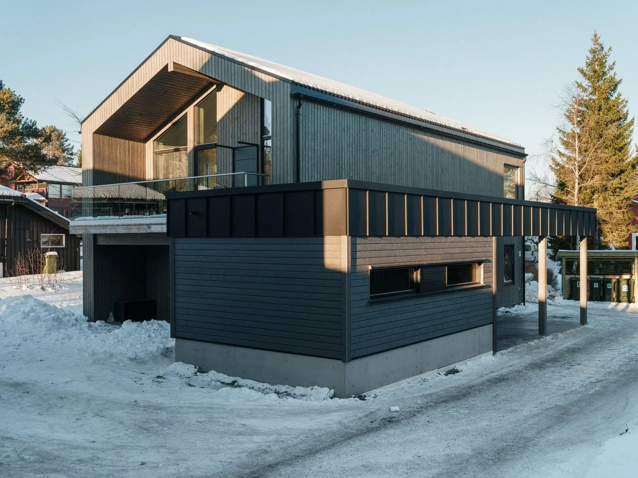 Modern two-story house with a gray and black exterior, large windows, and a balcony, situated in a snowy landscape with trees in the background.