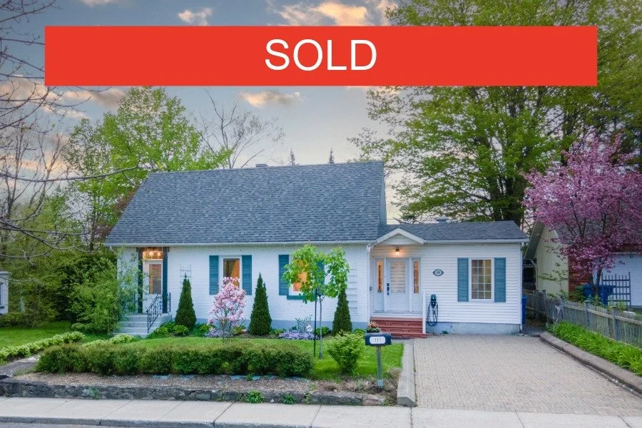 White house with blue shutters and a driveway, sold sign on top, trees and greenery surrounding the house.