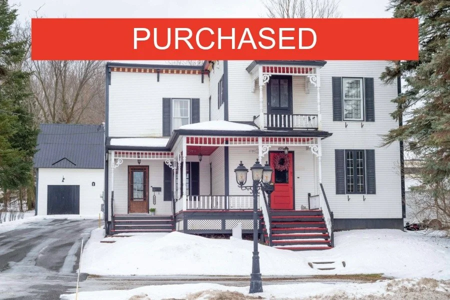 White multi-story house with black shutters and a red front door, snow on the ground, and a "PURCHASED" banner across the top.