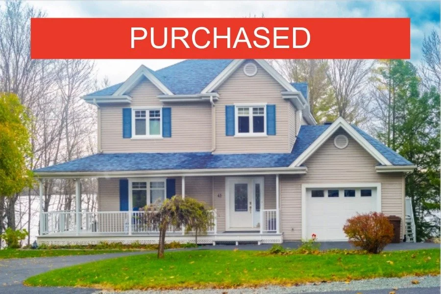 Two-story house with beige siding, white trim, and blue shutters, with a front porch and a white garage door, surrounded by trees and lawn, with a red "PURCHASED" banner at the top.