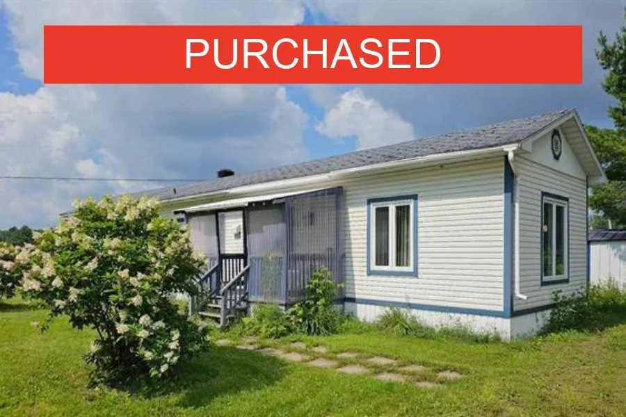 A house with white siding and blue trim, with a small porch and steps leading up to the entrance. There is a bush with white flowers, and the sky is partly cloudy. A red banner with white text reading "PURCHASED" is over the top of the image.