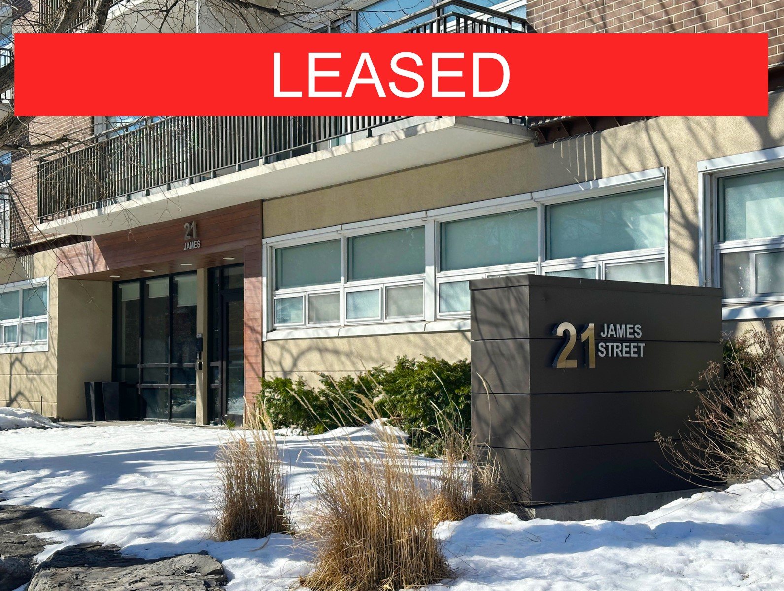 Apartment building with a sign reading '21 James Street' in front, snowy ground, bushes, and a large red banner with white text 'LEASED' at the top of the image.