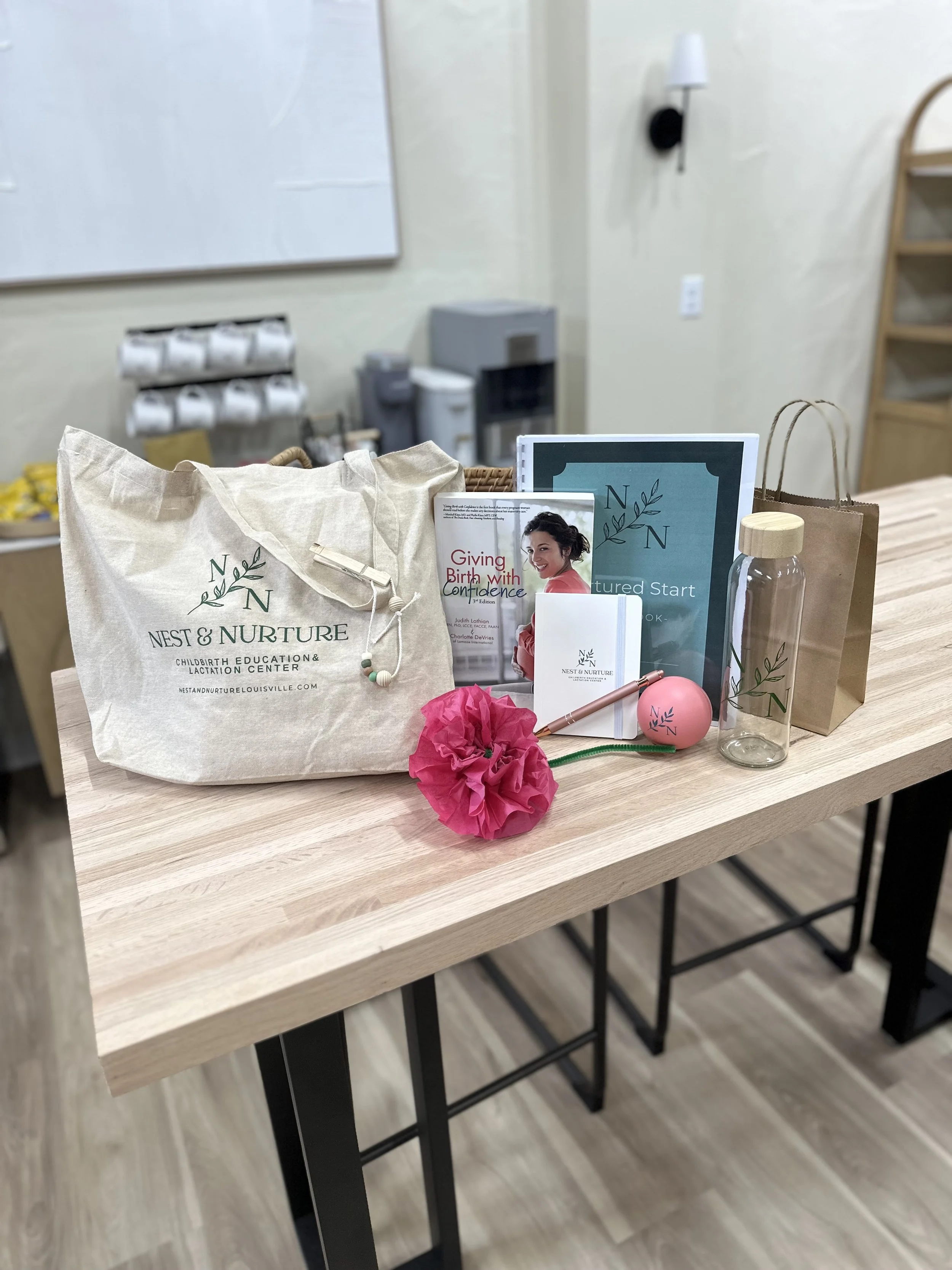 Display table with various items including books, a tote bag, water bottle, pink flower, and promotional materials from Nest & Nurture, a childbirth education and lactation center.