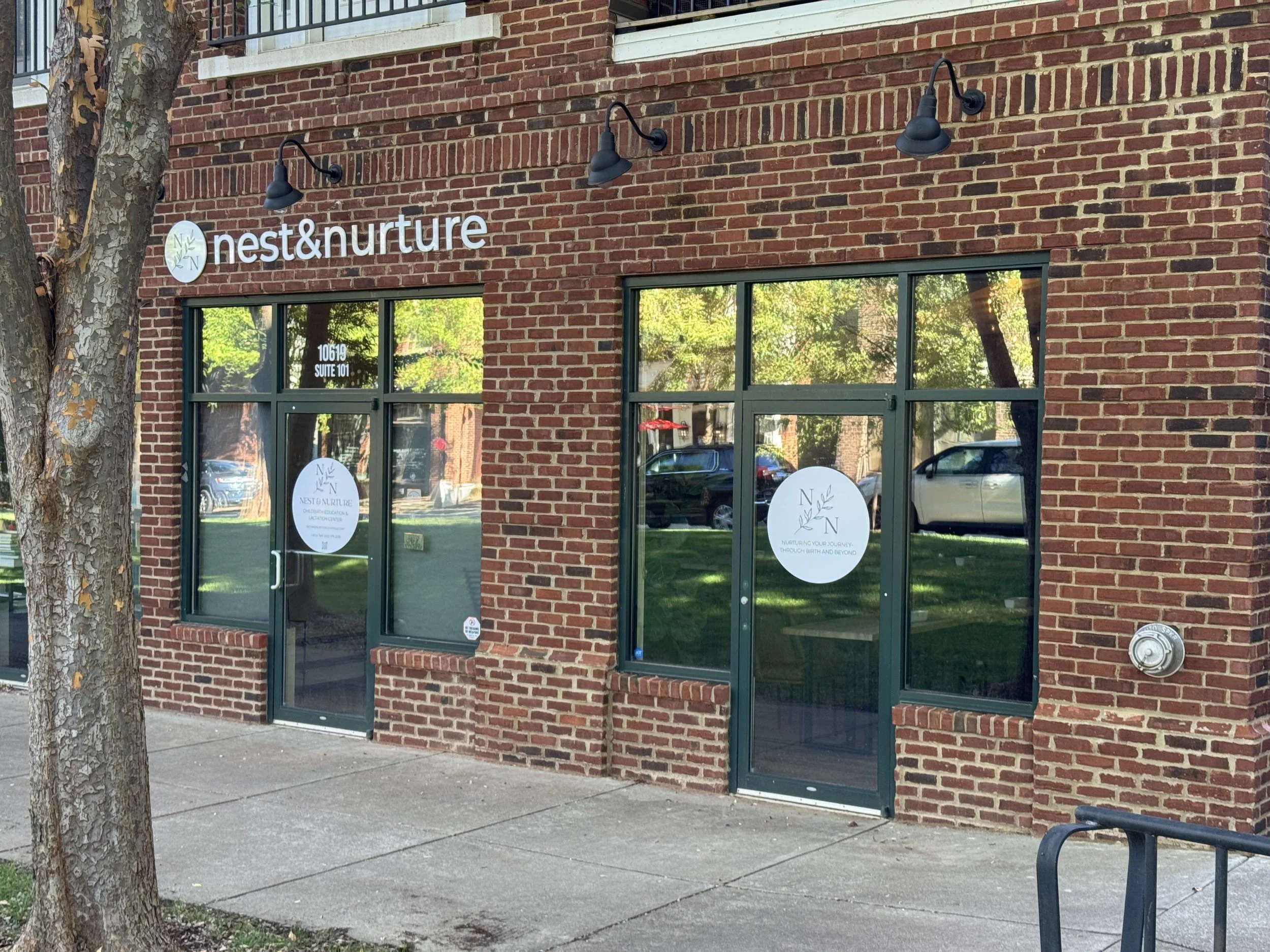 Brick storefront with large windows and a glass door, displaying signs for 'nest&nurture,' outdoor greenery reflected in the windows, and a sidewalk in front.
