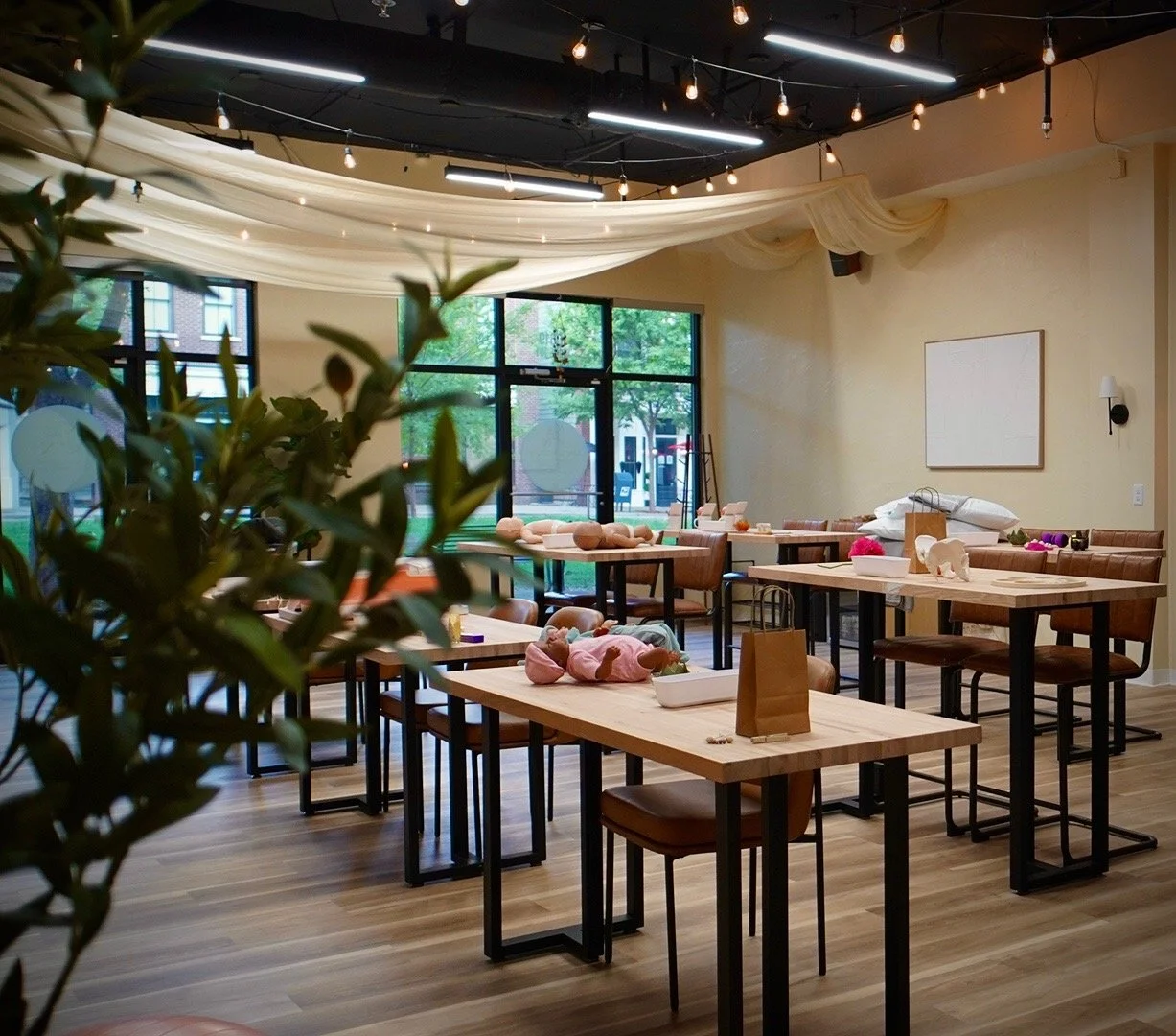 Empty restaurant or event space with wooden tables and chairs, some with stuffed animals, decorations, and paper bags. Large windows with greenery outside, string lights hanging from the ceiling, and draped fabric decor.