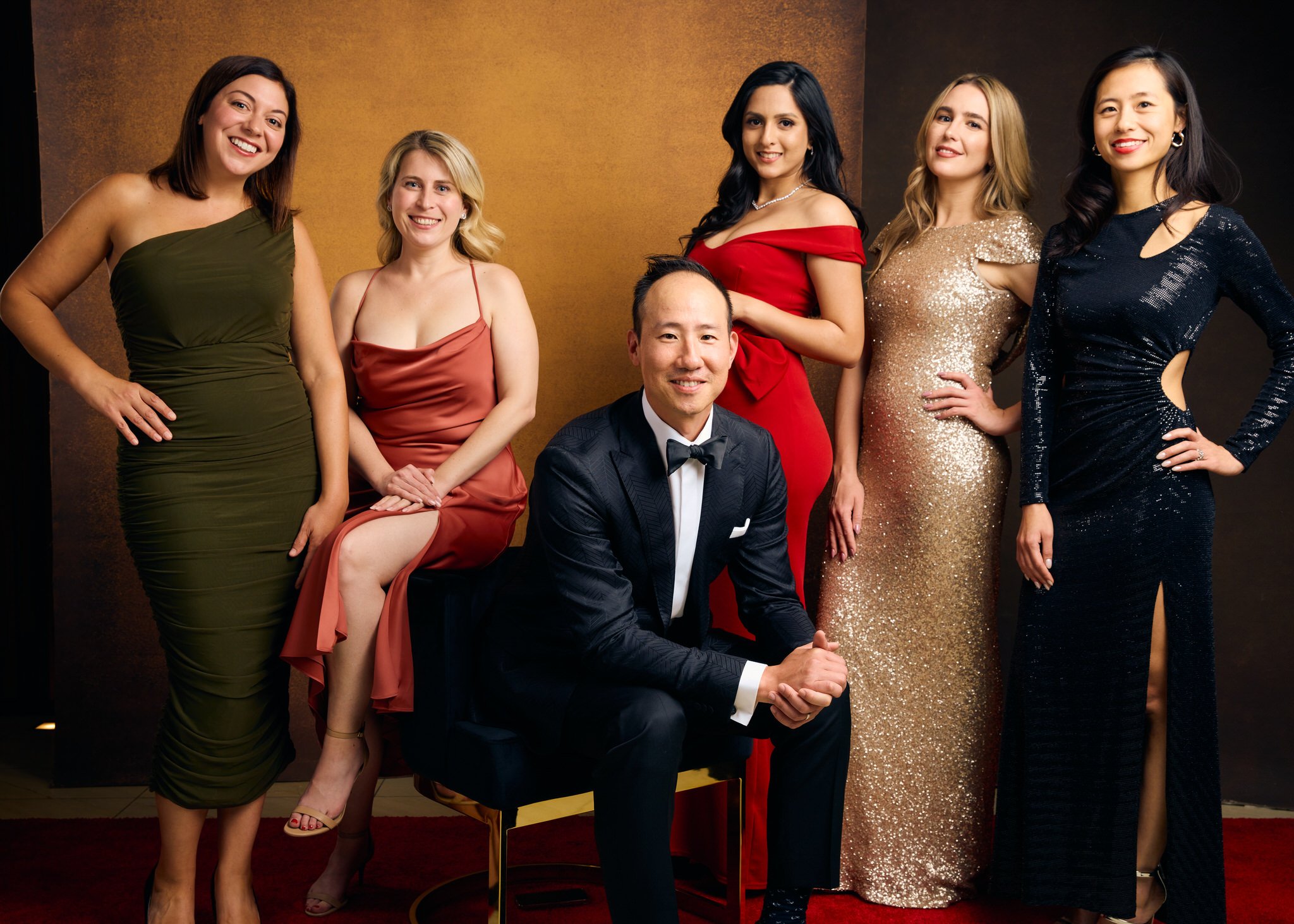 Six elegantly dressed gala guests pose for a polished Vanity Fair-style portrait against a warm studio backdrop.