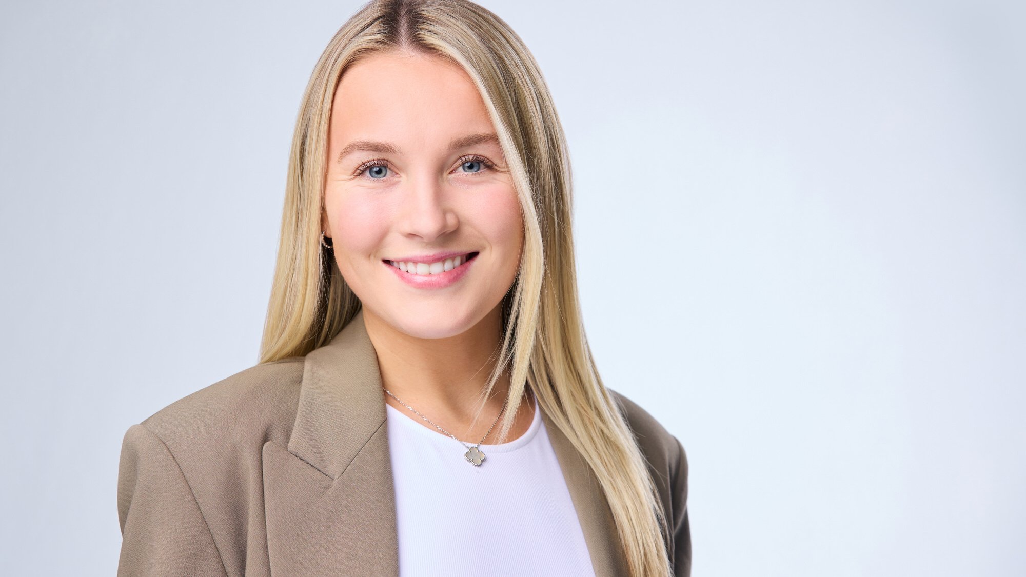 Professional headshot of a blonde woman in a beige blazer against a light blue background.