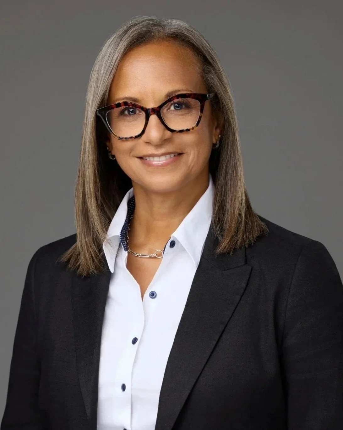 Professional corporate headshot of a woman in glasses and a dark blazer against a neutral grey studio background.