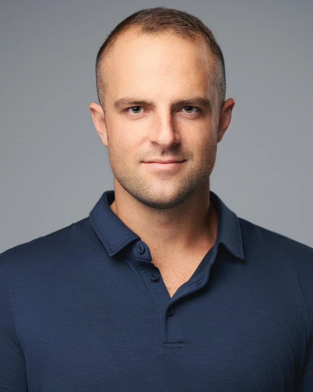Professional studio headshot of a man in a navy polo shirt against a dark grey background.