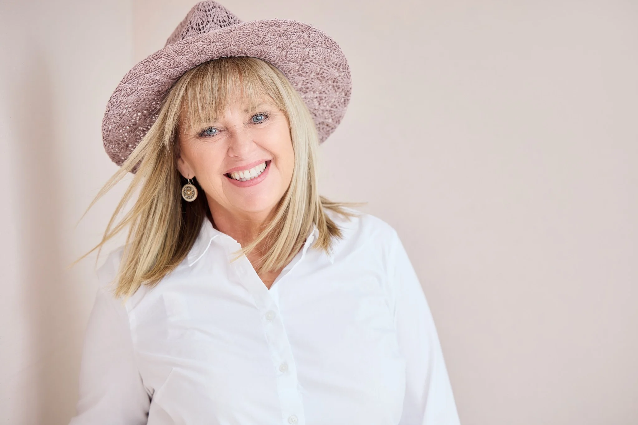 Cheerful personal branding headshot of a woman in a white top and hat against a soft pink studio background.